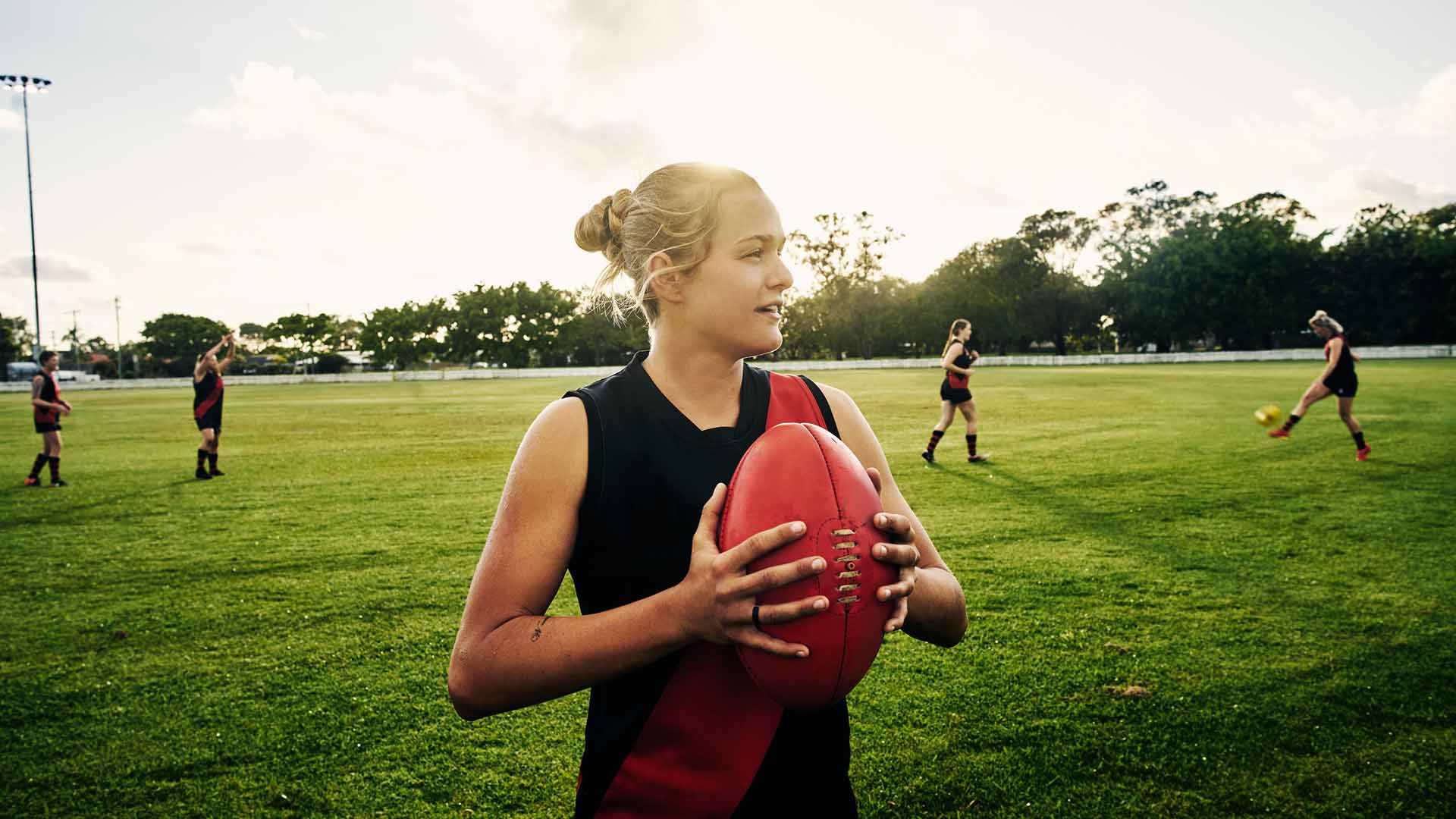 913653576 Shot of a young woman holding a football on a field with her team in the background