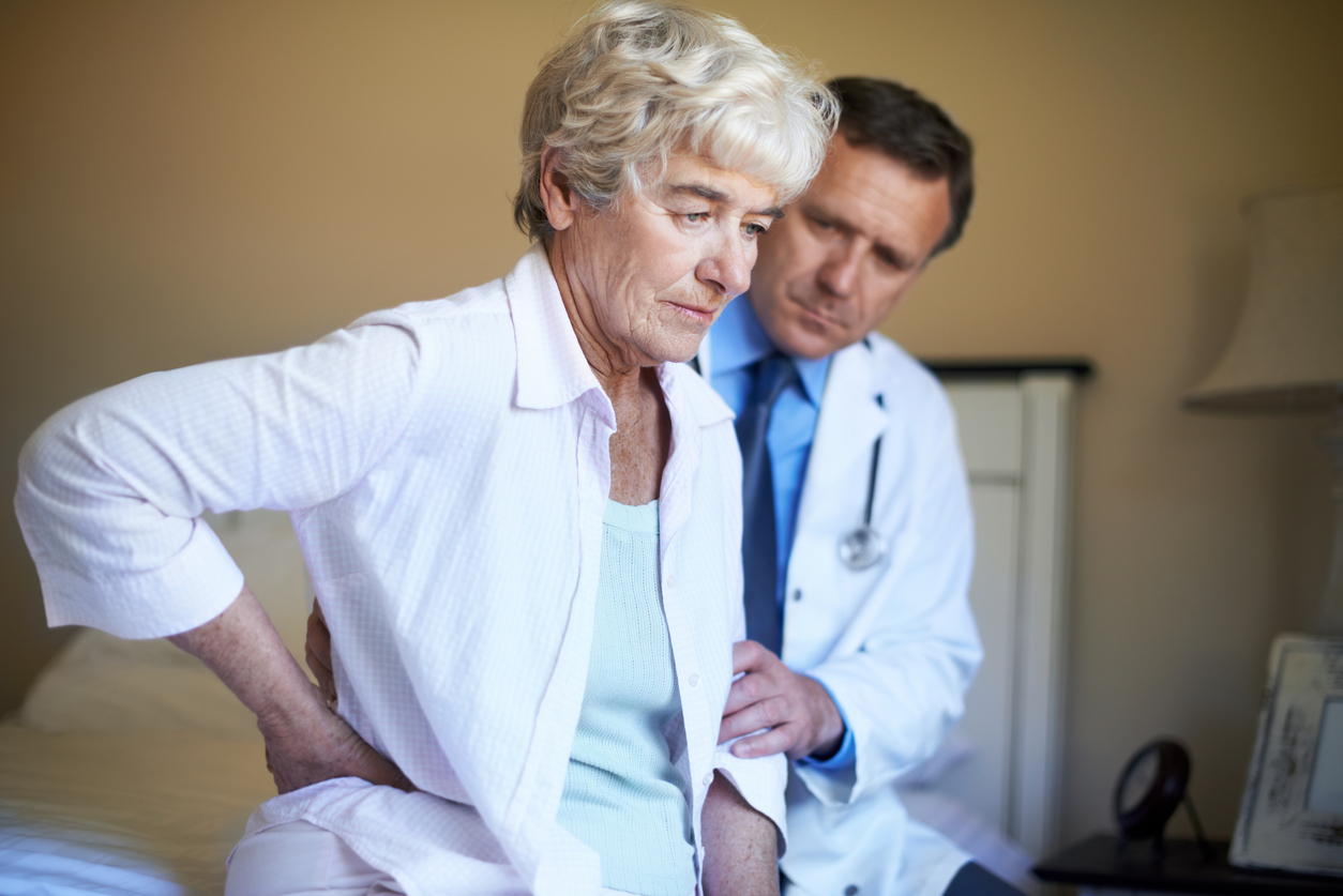 172462066 "Profile of a senior woman, sitting on her bed and holding her back in pain while her doctor sits besides her with a concerned expression on his face"