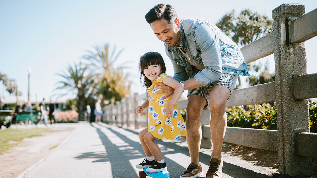 father helping young daughter to skateboard on a footpath father helping young daughter to skateboard on a footpath